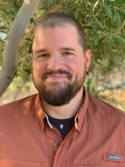 A smiling man with a beard wearing a rust-colored shirt outdoors.
