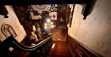 People gathered in a vintage, warmly lit hallway with a staircase.