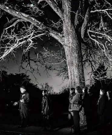 A man speaks to a group under a large tree at night in black and white.