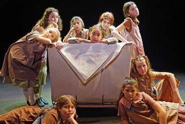 A group of nine girls in vintage dresses around a covered couch in a theatrical setting.