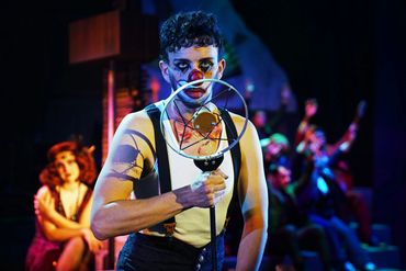 A performer with clown makeup holds a vintage microphone on stage.