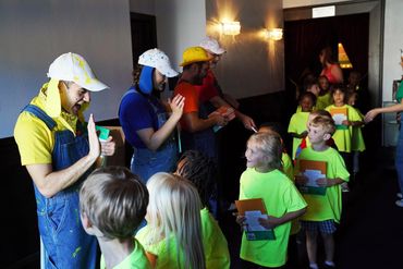 Adults in costumes greet children wearing bright yellow shirts holding books.