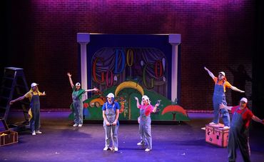 Actors in colorful overalls perform on stage with a vibrant backdrop.