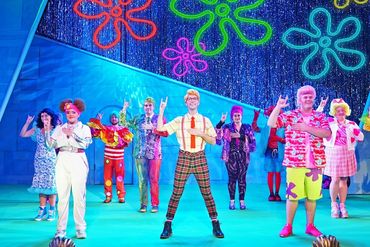 Cast of a colorful stage musical performing with neon flower lights in the background.