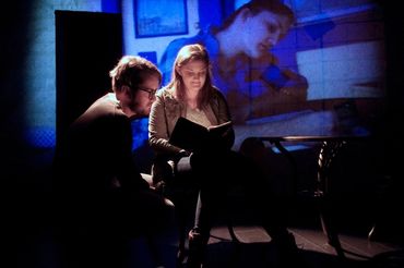 Two people read a book together under dim blue lighting with a projected image behind them.