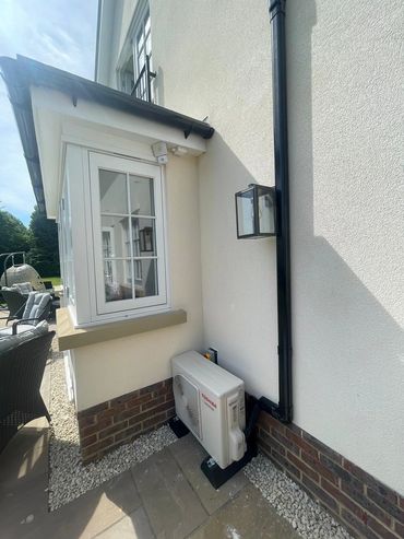 Outdoor air conditioning unit installed beside a house wall with patio furniture nearby.