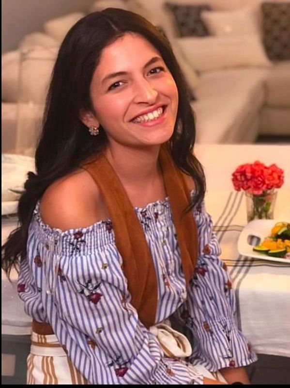 Smiling woman with long dark hair in a striped off-shoulder blouse sitting indoors.