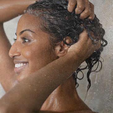 Mulher de pele morena, sorridente lavando os cabelos com shampoo natural, sem ingredientes sintético