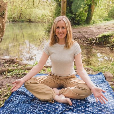 Woman sat in front of a lake on the ground, crossed legged with arms facing upwards.