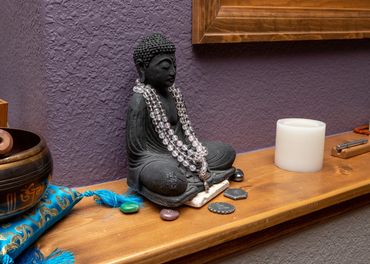 Buddha statue with beaded necklaces and stones on a wooden shelf.