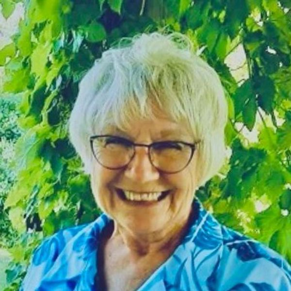Elderly woman smiling outdoors with green leafy background.