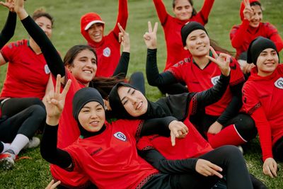 Women athletes in red sportswear posing happily on grass, showing peace signs.