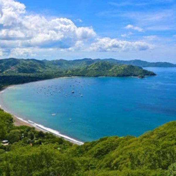 Scenic bay with clear blue waters, green hills, and scattered boats under a bright sky.