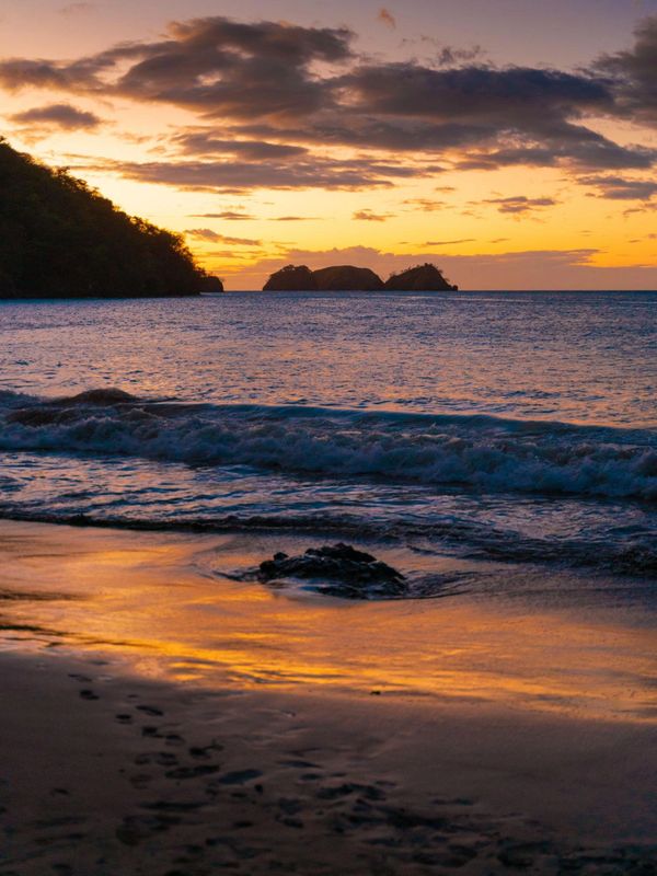 Sunset over a calm beach with gentle waves and distant islands.