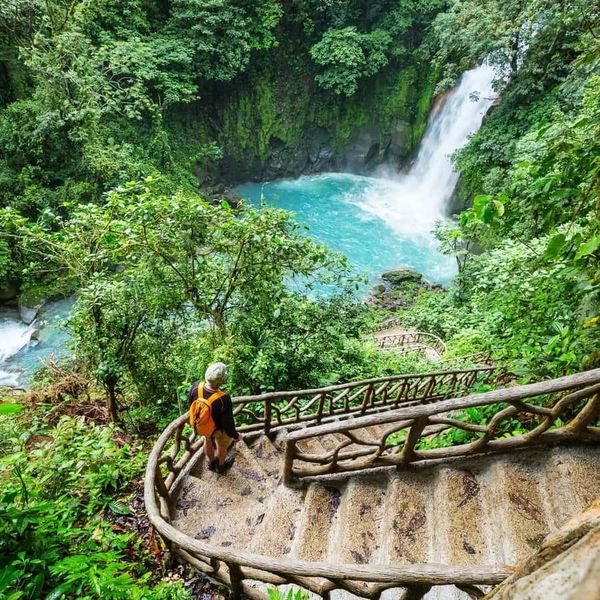 Person descending stairs toward a vibrant blue waterfall surrounded by lush greenery.