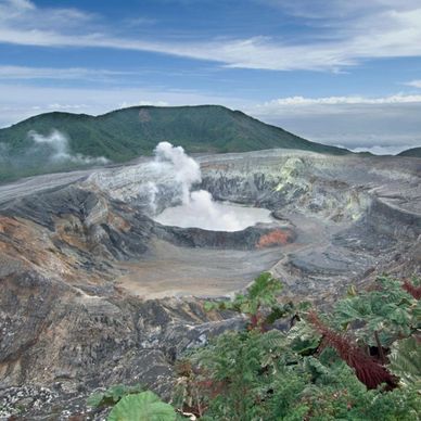 Volcanic crater emitting steam surrounded by greenery and mountains.