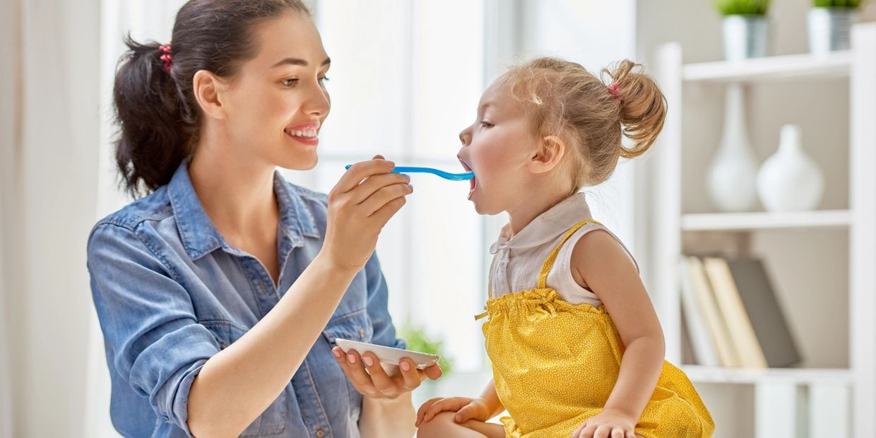 Mother smiles and feeds toddler with feeding problem with a spoon. Toddler leans forward to eat.
