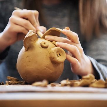 Hands sculpting a clay pig figurine with detail tools.