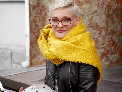 stylish woman with pixie cut, red glasses, black leather jacket and over sized yellow scarf smiling