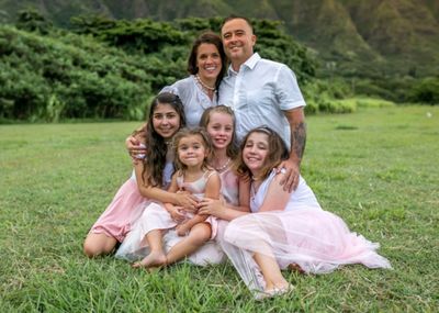 Smiling family of six in coordinated pastel outfits, posing outdoors on grass with greenery in the background.