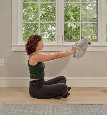 Woman sitting cross-legged indoors, stretching arms forward holding a cushion.