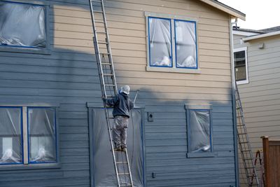 A painter on a ladder painting a house gray with windows covered in plastic.