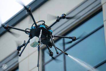 A drone sprays liquid in front of a building's glass windows.