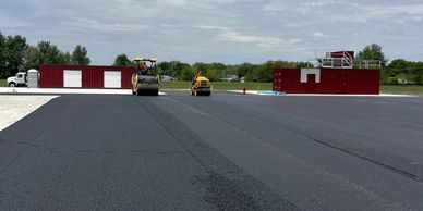 Two road rollers compacting freshly laid asphalt near red buildings under a partly cloudy sky.