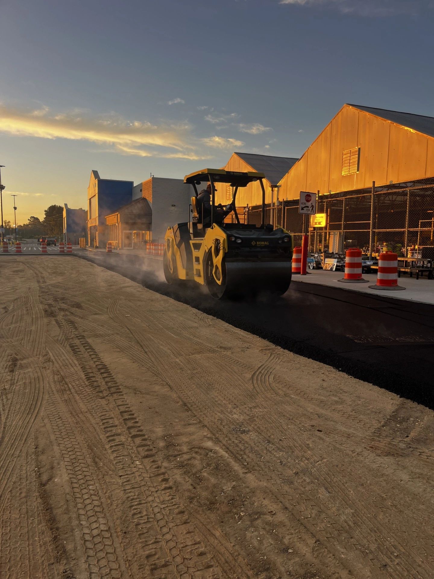 Road roller paving fresh asphalt near stores at sunset.