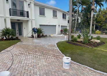 Two workers cleaning a brick driveway outside a large white house.