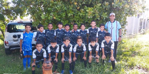 Youth soccer team posing outdoors with their coach beside a car under a tree.