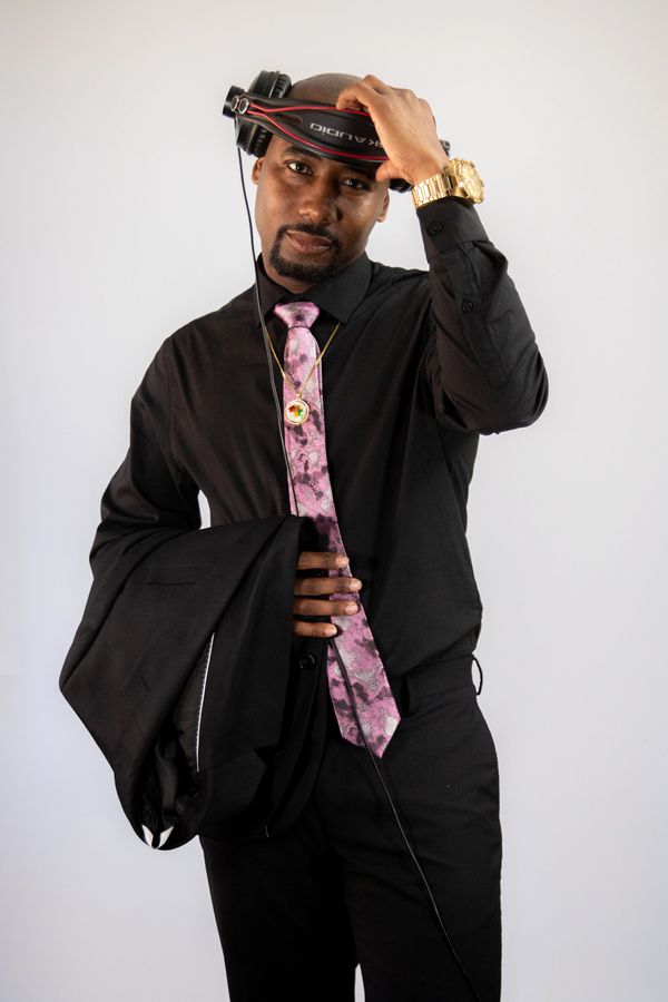 Man in black suit holding headphones and a jacket, wearing a pink tie and gold watch.