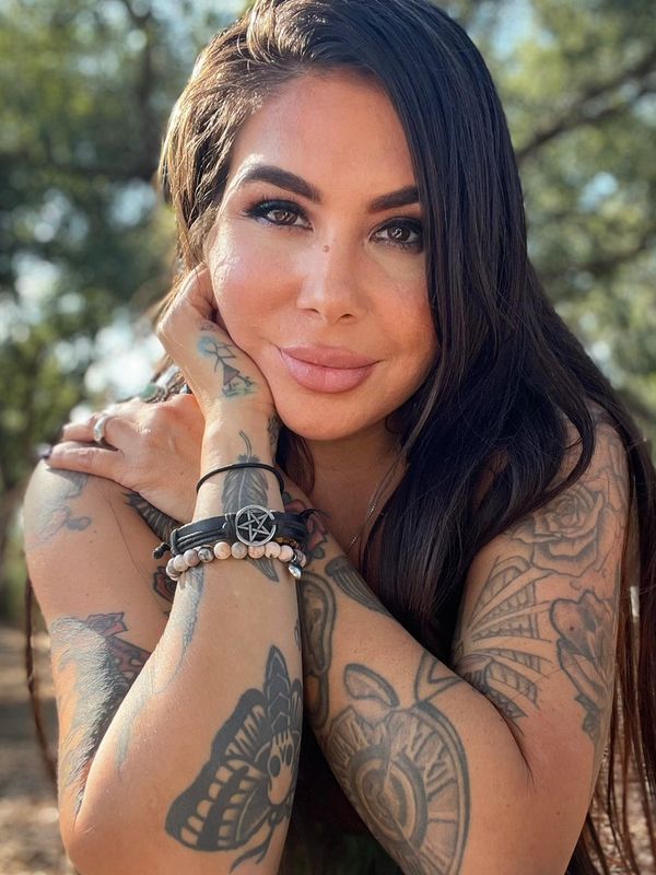 Smiling woman with long dark hair and detailed tattoos on her arms in an outdoor setting.