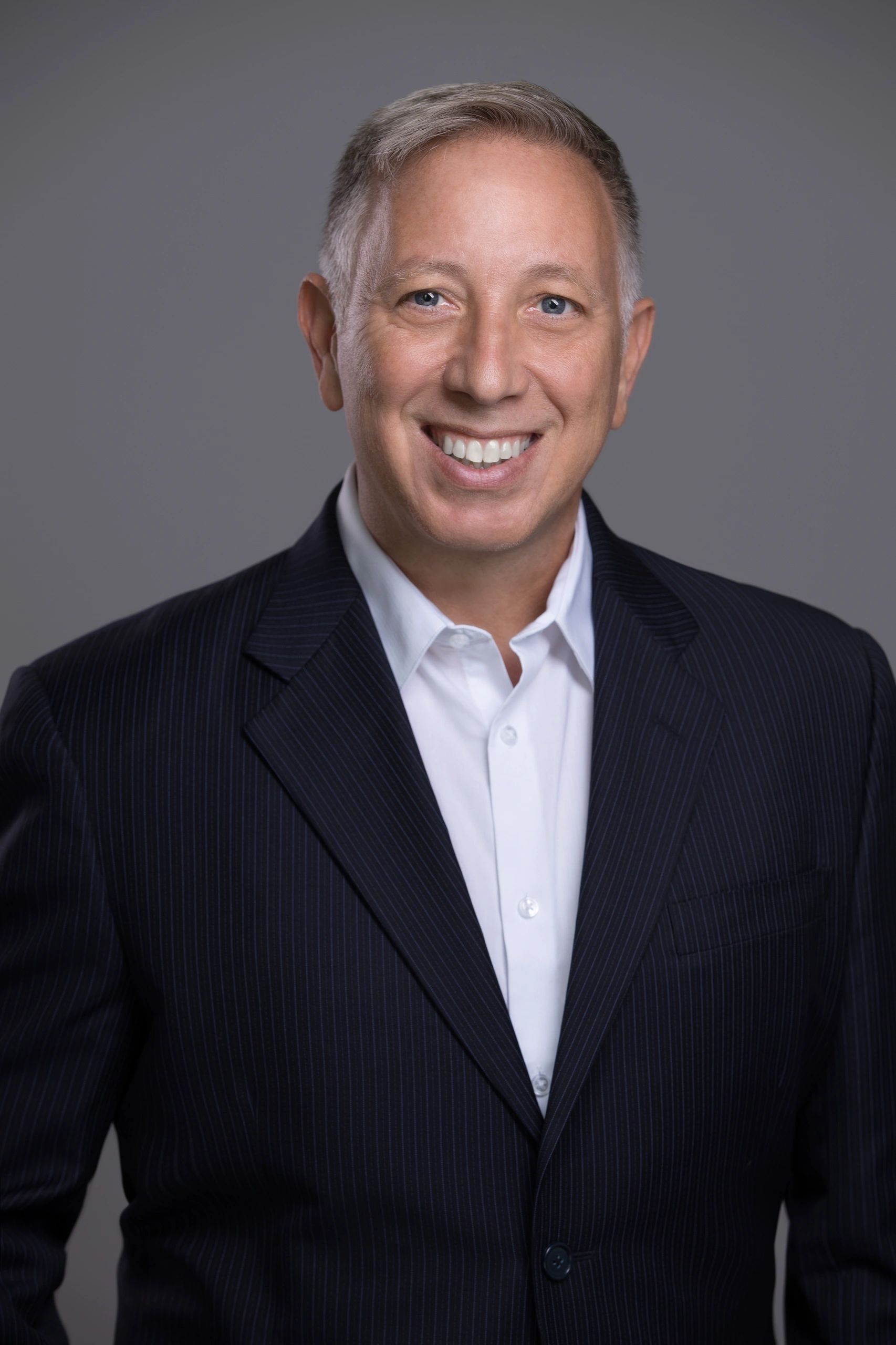Smiling man in a dark pinstripe suit and white shirt against a gray background.