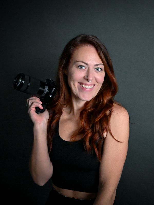 Smiling woman with red hair holding a camera against a dark background.