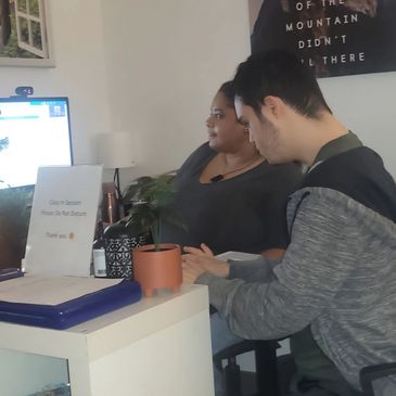 Two people sitting side by side at a desk with a computer and plant.