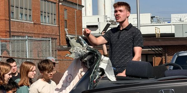 Young man speaks energetically beside a damaged car roof with attentive onlookers.