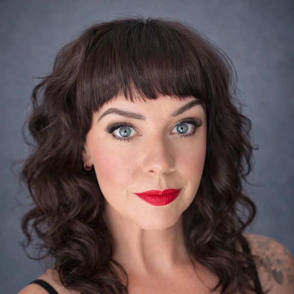 Woman with curly dark hair and bright red lipstick posing against a gray background.
