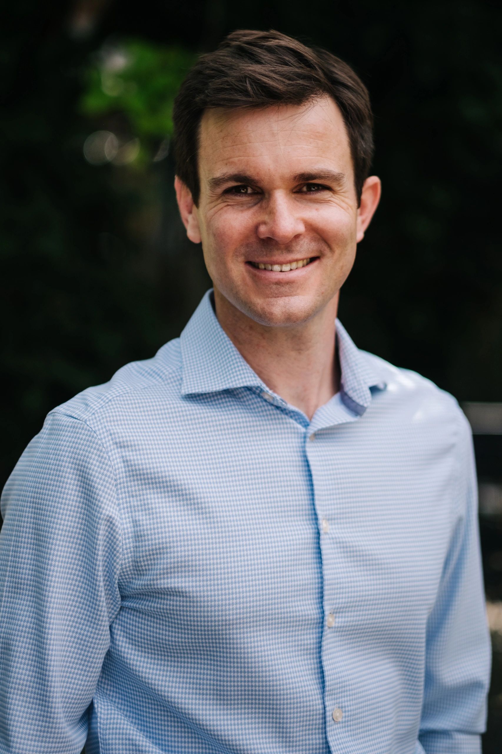 Smiling man in a light blue checkered shirt outdoors.