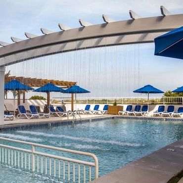Outdoor swimming pool with blue lounge chairs and umbrellas under a pergola waterfall feature.