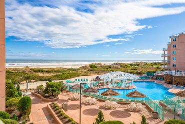 Sunny beachside pool area with lounge chairs and umbrellas near the ocean.