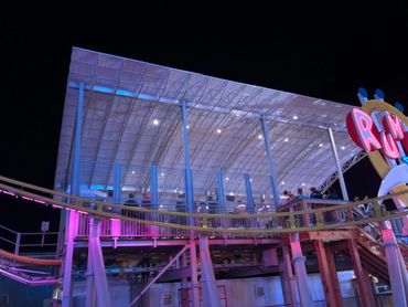 People wait in line on a lit-up roller coaster platform at night.