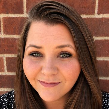 Smiling woman with long brown hair standing against a brick wall.