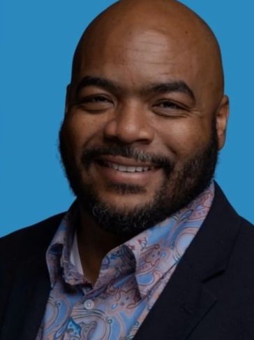 Smiling man with a beard wearing a patterned shirt and black blazer against a blue background.