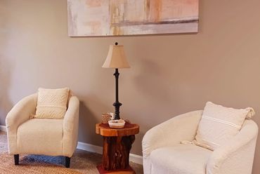 Two beige armchairs with cushions flank a wooden side table and lamp against a plain wall.