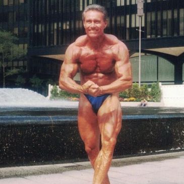 Muscular man posing in blue trunks outdoors near a fountain.