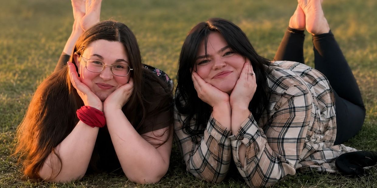 Two smiling women lying on grass, resting their faces on their hands.