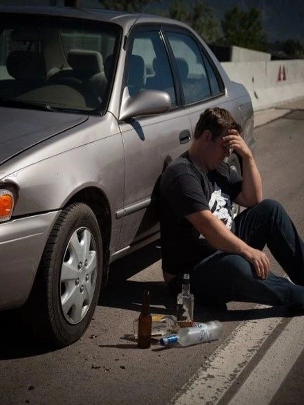 Man sitting on the road beside a car with empty alcohol bottles looking distressed.