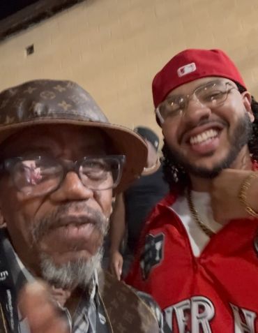 Two men smiling, one in a red cap and jacket, the other in a patterned hat and glasses.