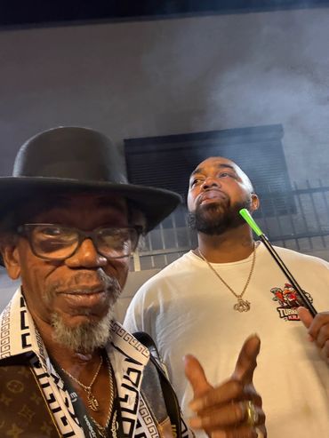 Two men posing, one wearing glasses and hat, the other smoking with a green pipe.